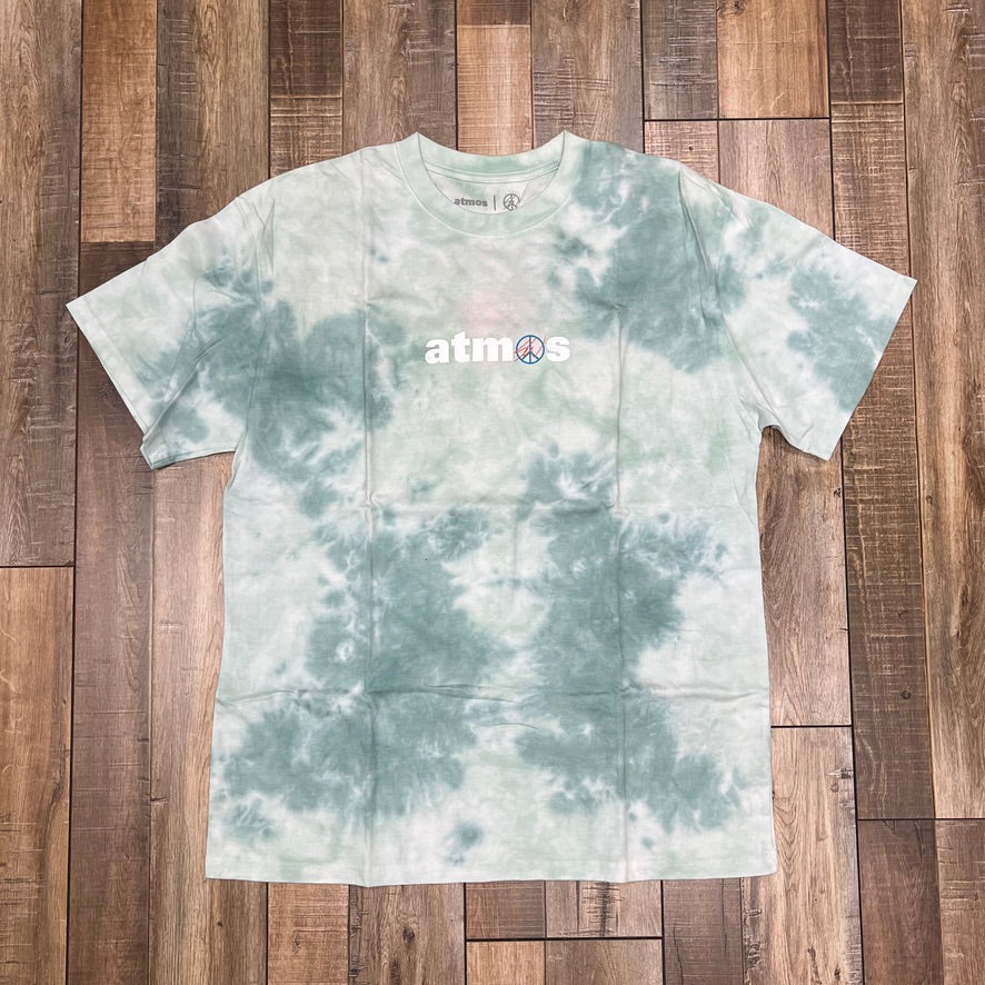 A brand new Atmos Sean Wotherspoon SW Green Tie Dye T-Shirt from Holy Ground Sneaker Shop features atmos in white with a peace sign as the o, displayed flat on a wooden floor.