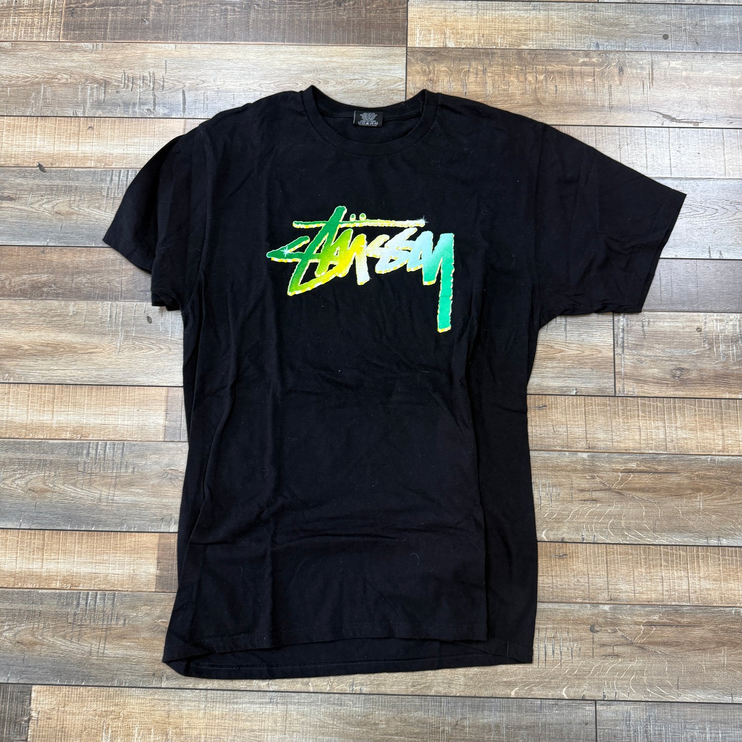 Laid flat on a wooden floor, this preowned Stussy Chrome T-Shirt features a stylized logo in a yellow, green, and white gradient across the chest.