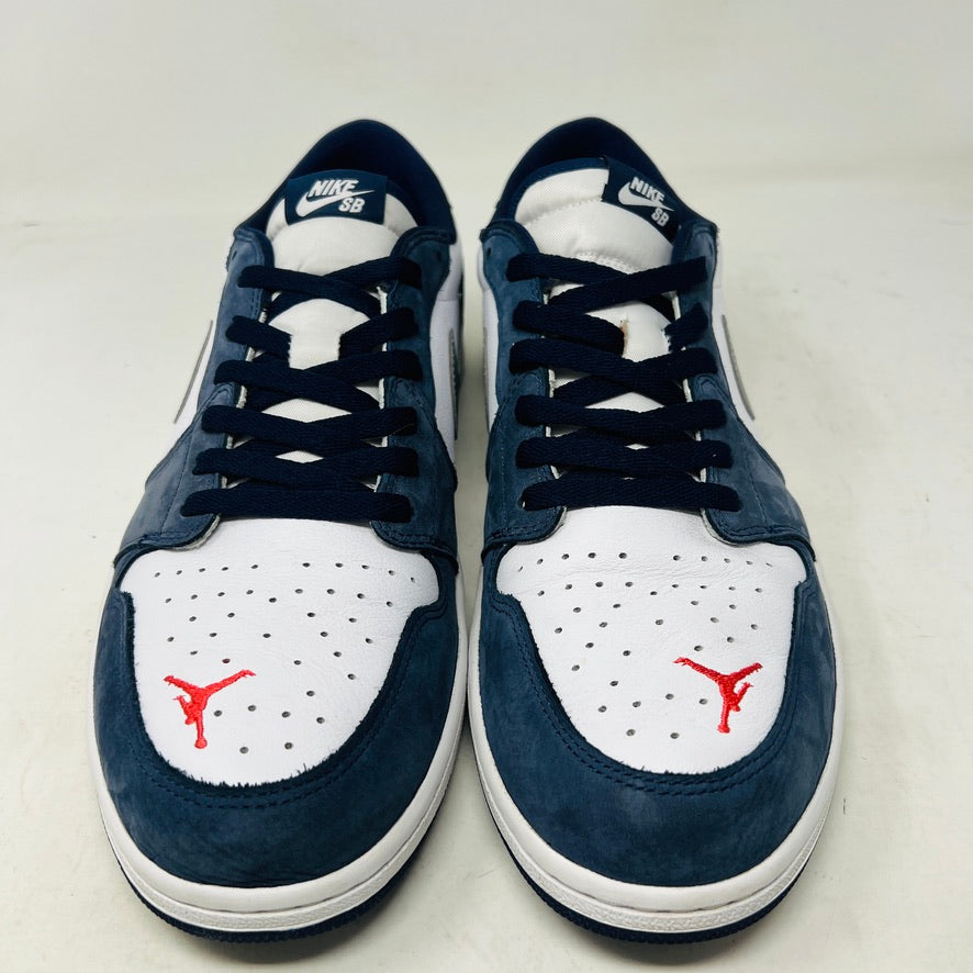 The Jordan 1 Low SB Midnight Navy sneakers, with white and navy panels and a side Nike swoosh, are displayed on a pristine shoebox. A spare set of white laces featuring the Nike SB logo in Metallic Silver is also included.