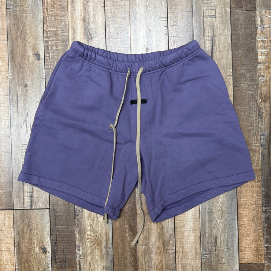 A preowned pair of Essentials Heavy Fleece Short Lavender is laid flat on a wooden floor, featuring a drawstring waistband, two side pockets, a small black tag below the waistband, and soft-looking fabric.