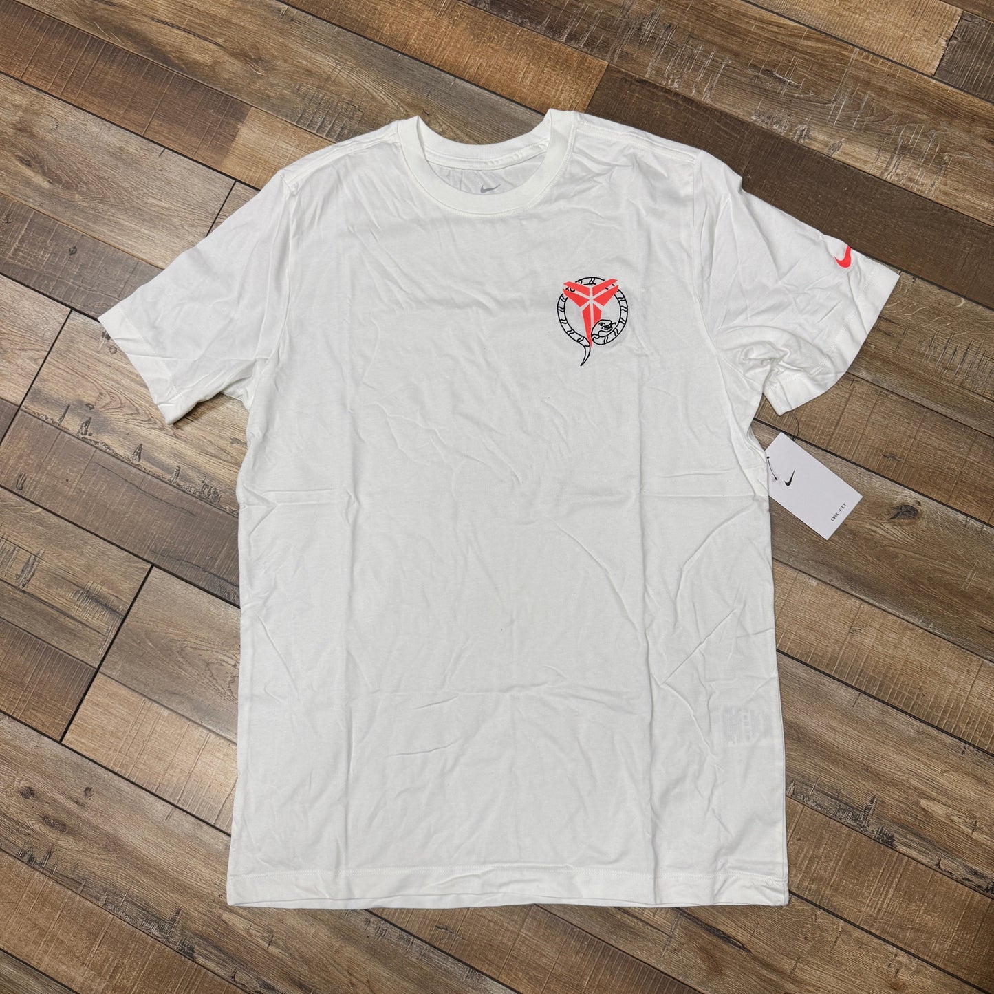 The Nike Kobe Dri-FIT Tee Stockings White features a heart-shaped snake and wings graphic on the left chest. A Nike logo adorns the left sleeve, with a tag attached, all displayed on a wooden floor.