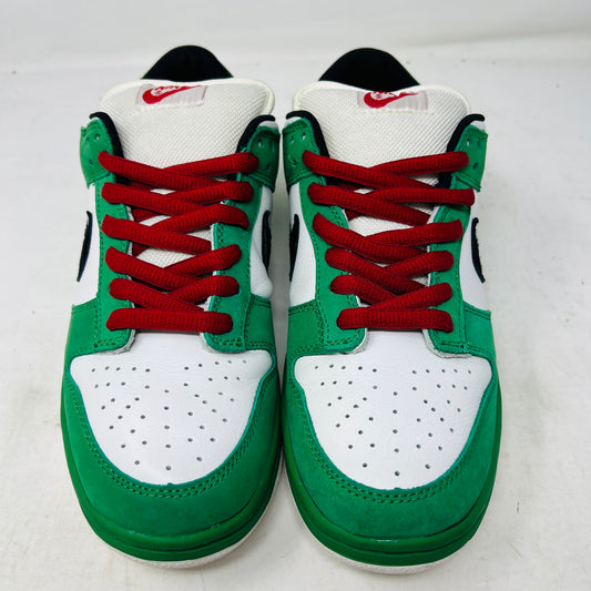 The Nike SB Dunk Low Heineken sneaker, featuring a green, white, and black design with red laces and a red star on the heel, sits on its box. A small package of green shoelaces is beside it. The box displays the size and Nike branding.