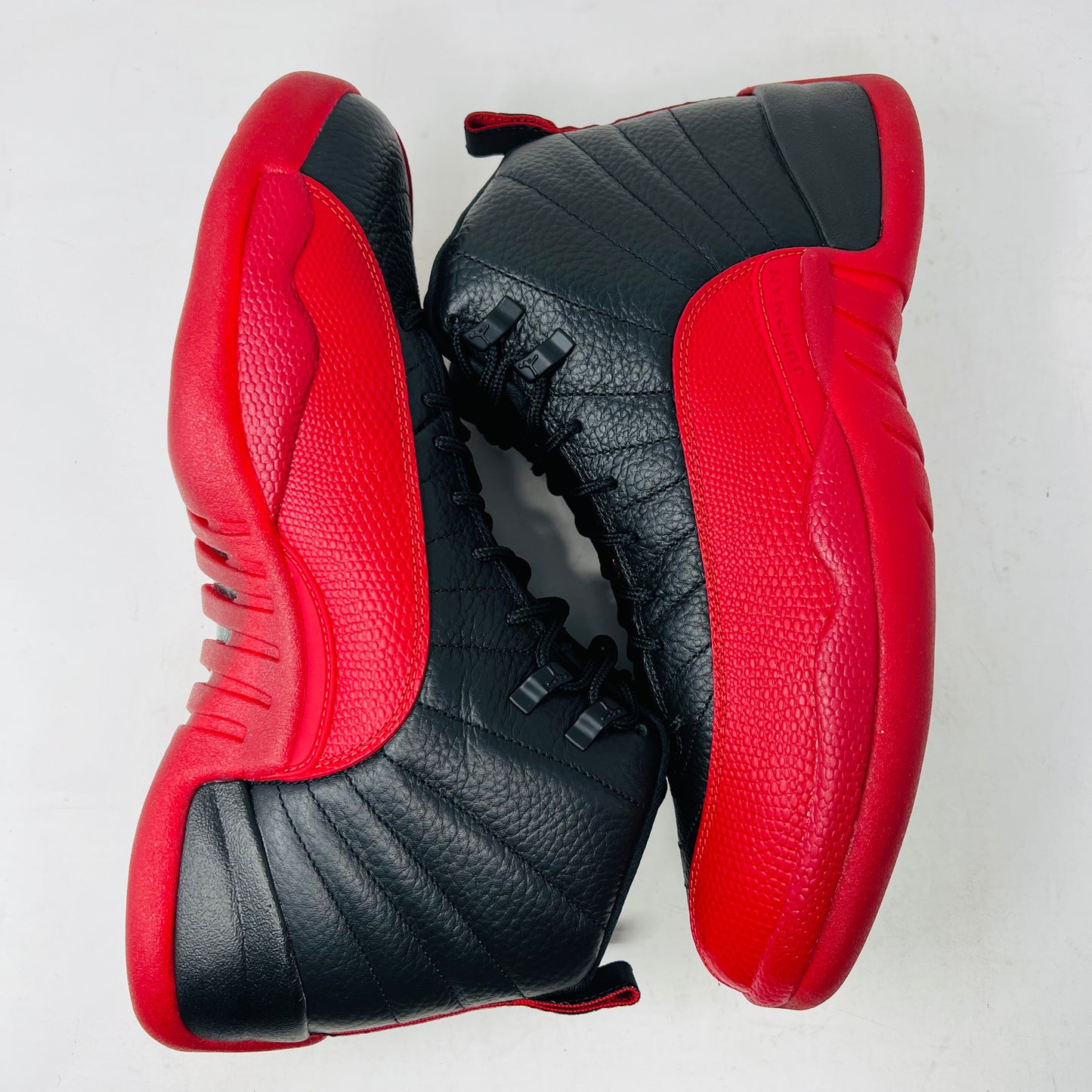 The Jordan 12 Retro Flu Game (2025) athletic shoe in black and red, brand new and pristine, sits atop an immaculate orange-and-black Nike box. It features a textured design with JUMPMAN embossed on its high-top style, perfectly enhancing this stylish accessory from the Jordan brand.