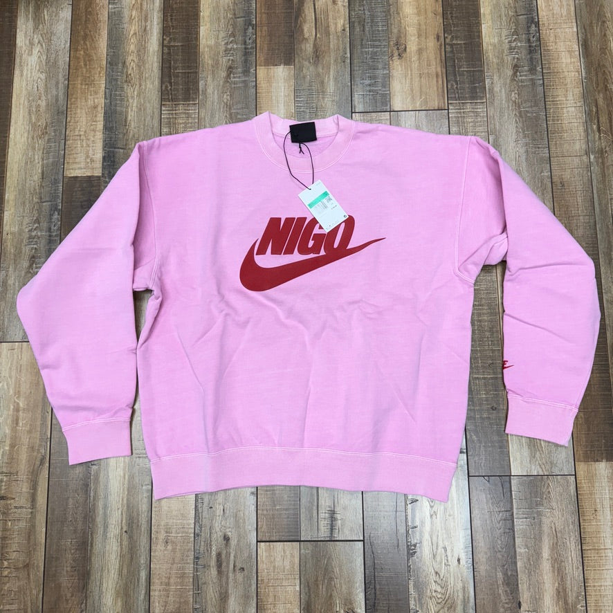 Brand new Nike x Nigo NRG Fleece Crew Sweatshirt in pink, featuring a red curved logo and Nigo text on the front, with tag attached. Laid flat on a wooden floor.