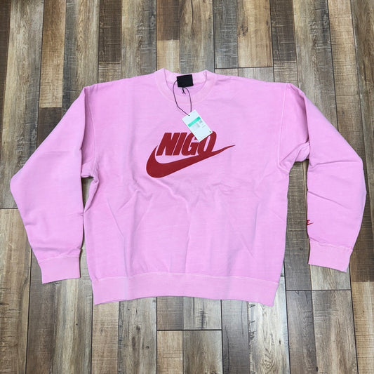 Brand new Nike x Nigo NRG Fleece Crew Sweatshirt in pink, featuring a red curved logo and Nigo text on the front, with tag attached. Laid flat on a wooden floor.