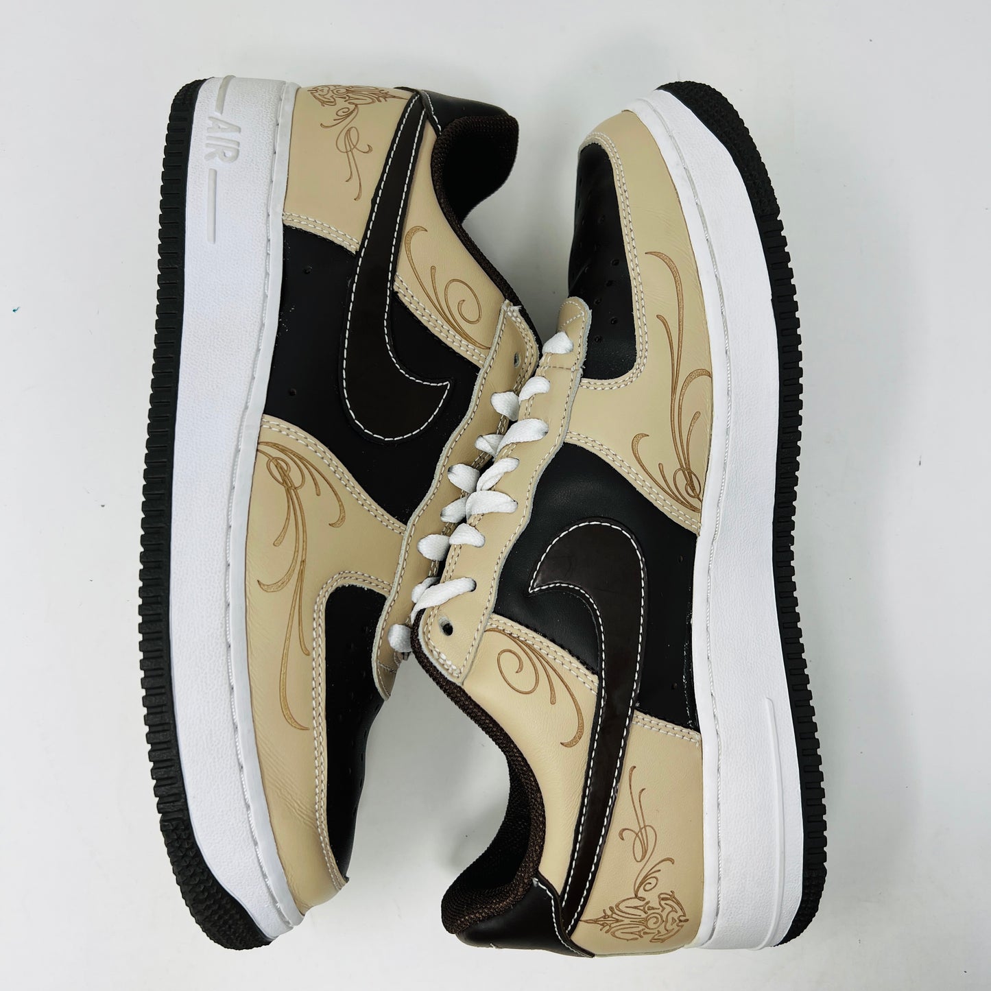 The Nike Air Force 1 Low Brown Pride sneakers feature beige and dark brown leather panels, white laces, and a white sole. They are shown from a top-down perspective in super clean condition on an immaculate white background.