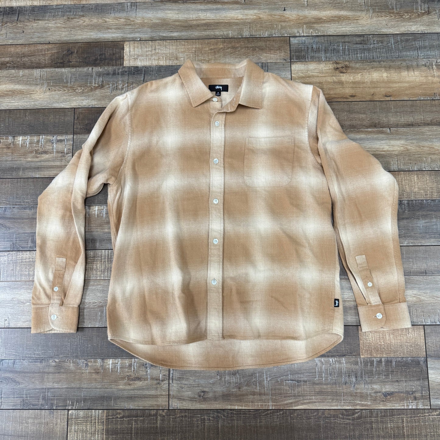 The Stussy Bay Plaid Shirt in Tan, by Stussy, is displayed on a wooden floor. It features long sleeves, white buttons, and a pointed collar with a soft beige and white plaid pattern.
