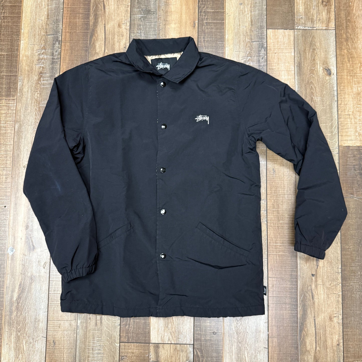 Black preowned Stussy Coaches Jacket on wooden floor.