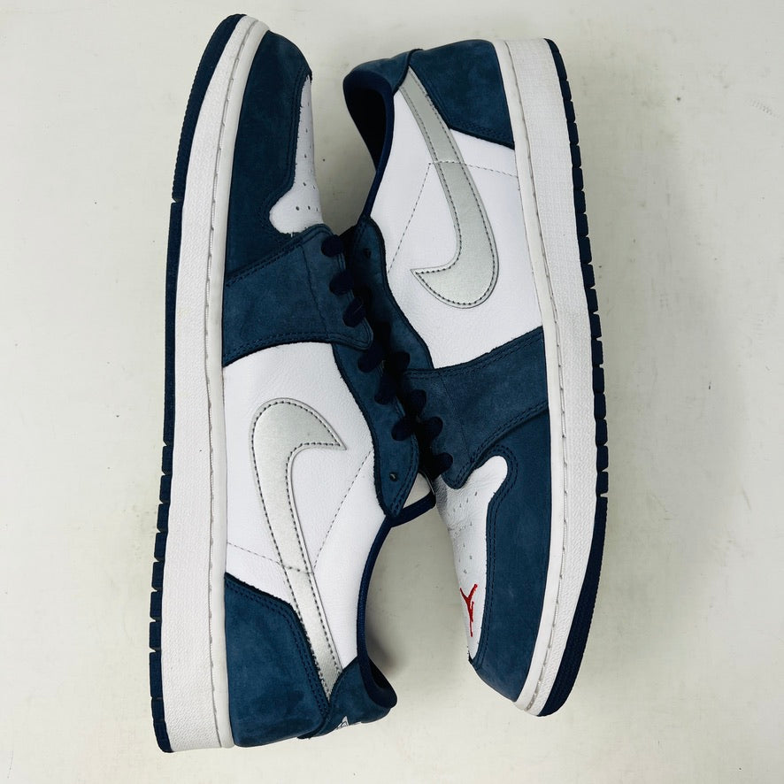The Jordan 1 Low SB Midnight Navy sneakers, with white and navy panels and a side Nike swoosh, are displayed on a pristine shoebox. A spare set of white laces featuring the Nike SB logo in Metallic Silver is also included.