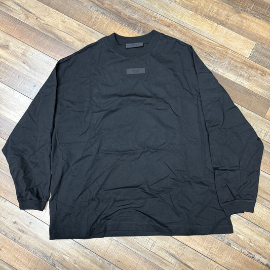 A pristine Fear of God Essentials L/S T-shirt in black, featuring a textured logo patch, is displayed on the wooden floor. A few natural wrinkles add character to its condition.