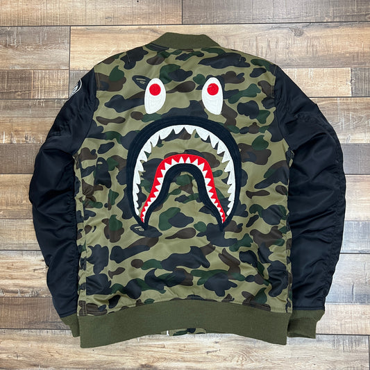 The preowned BAPE Shark MA-1 Jacket by Bape in green and brown camo features a black sleeve with the embroidered WGM letters and a cartoon tiger design, all displayed on a wooden floor.