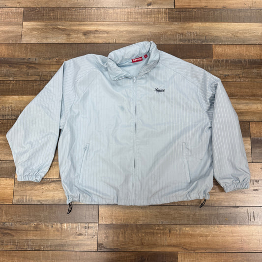 A Supreme Reflective Pinstripe Track Jacket in light blue, featuring a zippered front, two side pockets, elastic cuffs, and a small chest logo is laid flat on a wooden floor.