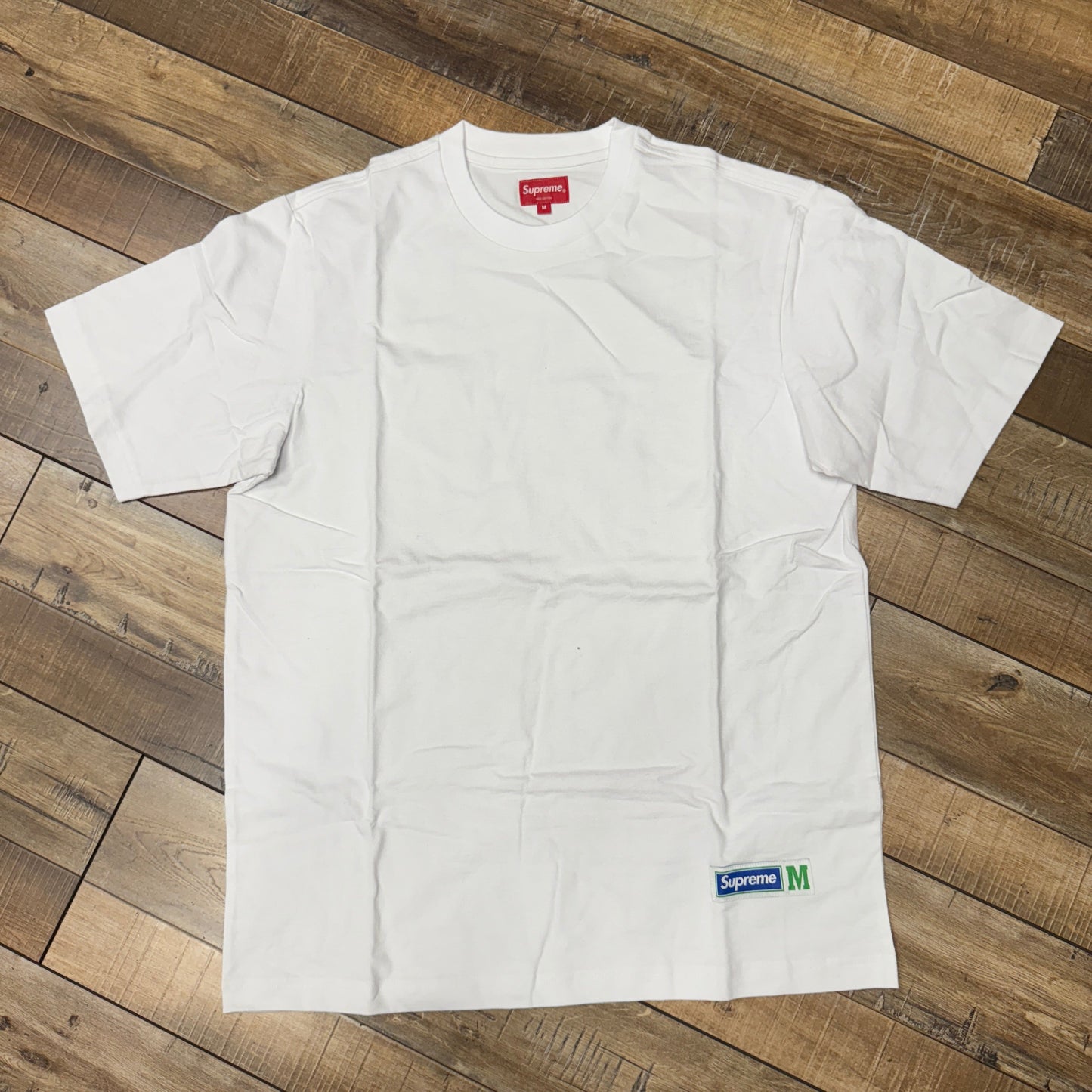 The Supreme Athletic Label S/S Top White is displayed on a wooden floor, featuring a red Supreme tag inside the collar and a small green and white Supreme label with M on the bottom edge, adding distinct elegance to this impeccable product.