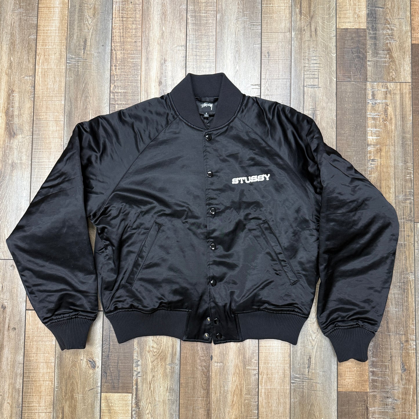 The Stussy Souvenir Jacket FW24 by Holy Ground Sneaker Shop is a black satin bomber with ribbed cuffs, collar, and hem. It features a snap button front and Stussy embroidered in white on the left chest. The jacket is laid flat on a wooden floor.