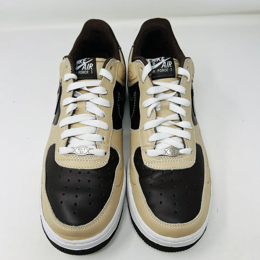 The Nike Air Force 1 Low Brown Pride sneakers feature beige and dark brown leather panels, white laces, and a white sole. They are shown from a top-down perspective in super clean condition on an immaculate white background.