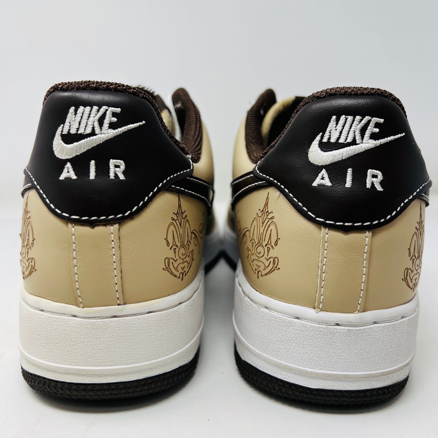 The Nike Air Force 1 Low Brown Pride sneakers feature beige and dark brown leather panels, white laces, and a white sole. They are shown from a top-down perspective in super clean condition on an immaculate white background.