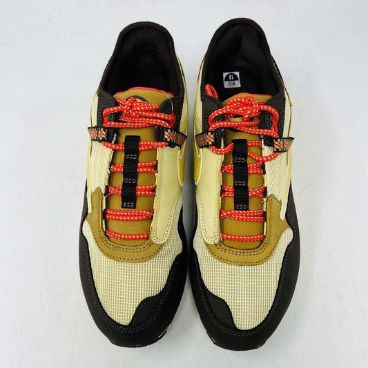 A brand new Nike Air Max 1 TS Cactus Jack Baroque Brown sneaker in yellow and brown with orange accents sits on a brown Nike shoebox. It boasts a wavy design, textured fabric, extra brown laces, and a visible air unit in the sole against a plain white background.