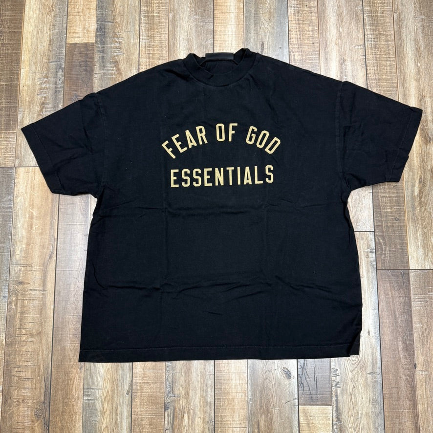 Preowned Essentials Fear of God black jersey crewneck tee with tan logo, displayed on a wooden floor.