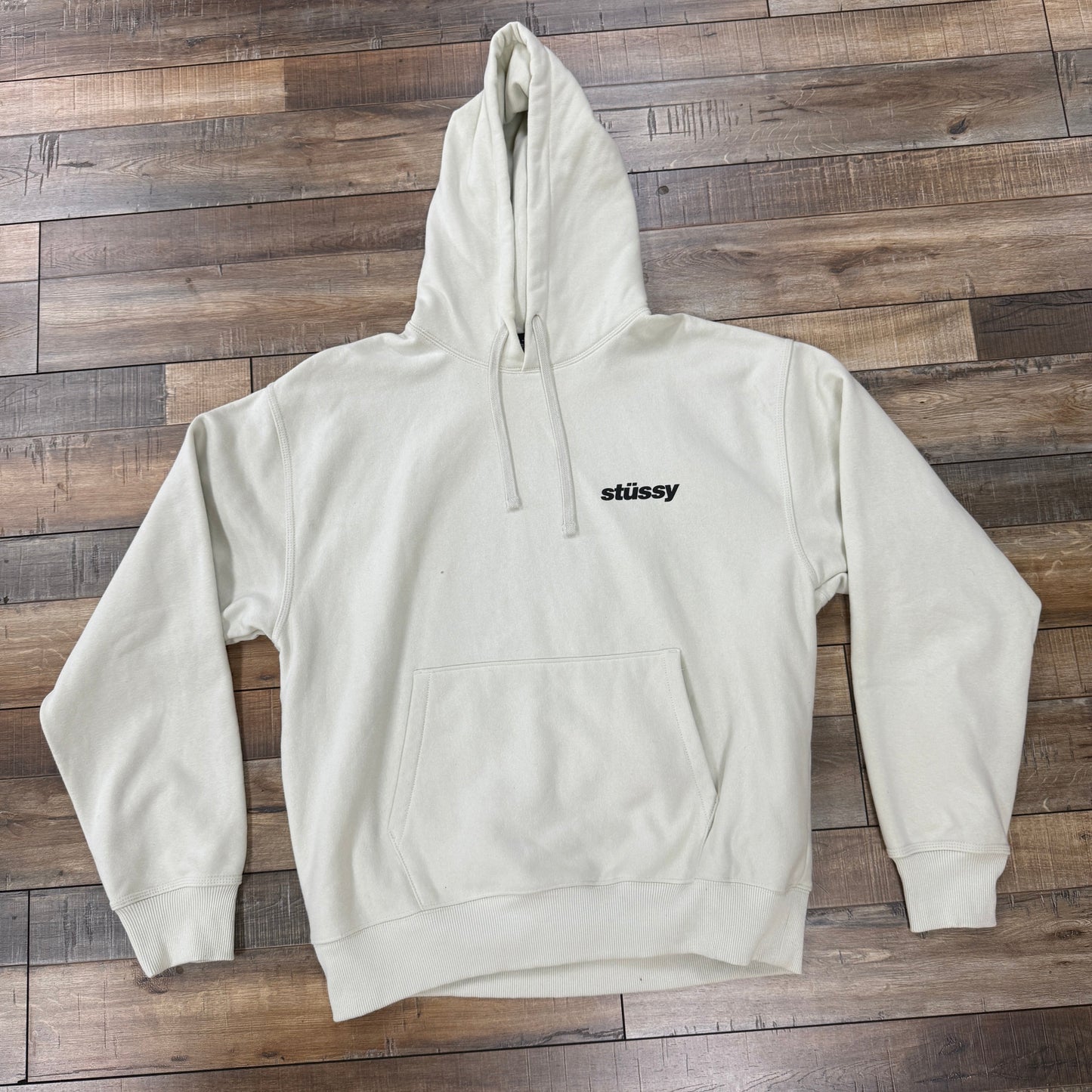 A preowned Stussy Popsicle Hoodie Stone in light beige, featuring a front pocket and Stussy in black text on the left chest, is displayed on a wooden floor with a mix of light and dark brown tones.