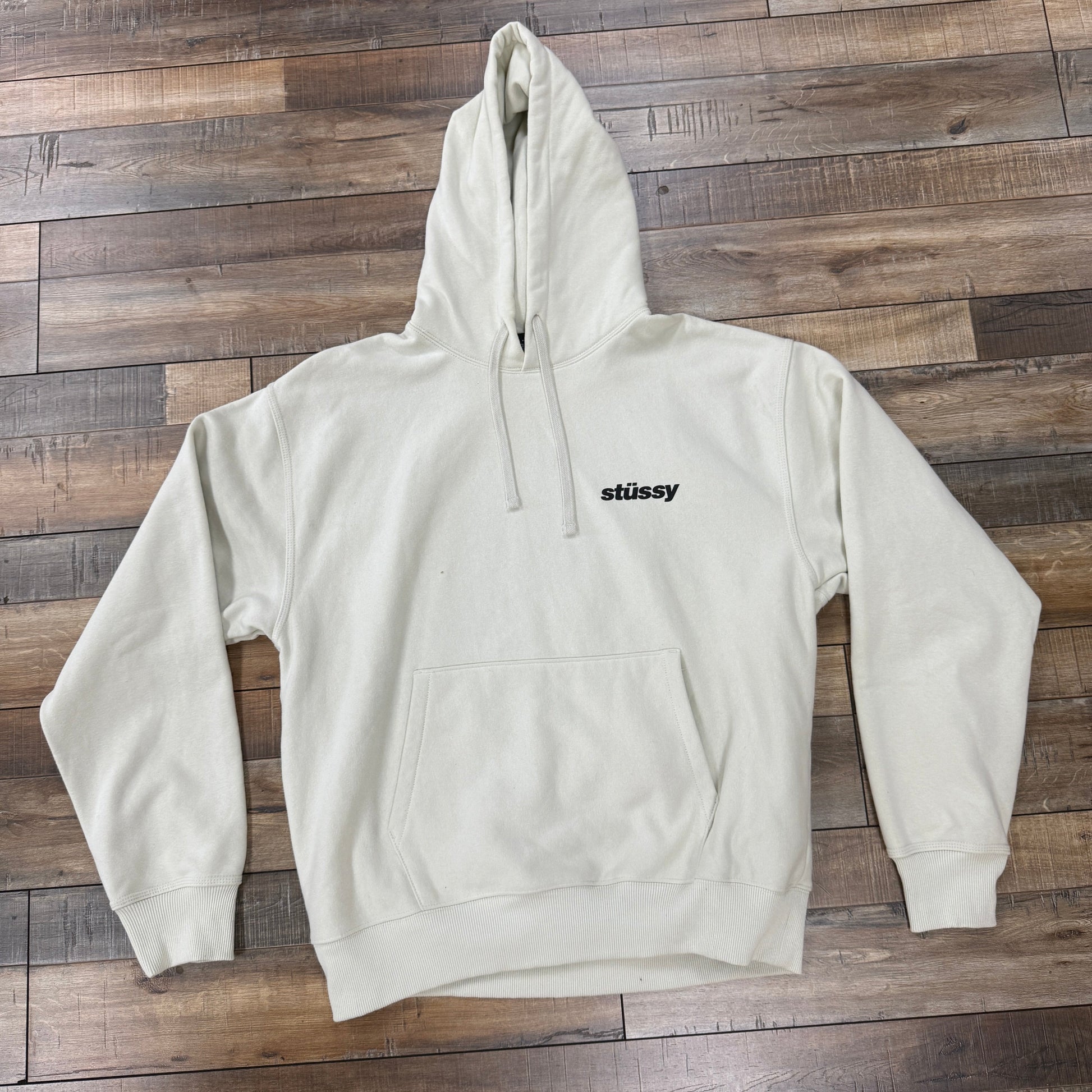 A preowned Stussy Popsicle Hoodie Stone in light beige, featuring a front pocket and Stussy in black text on the left chest, is displayed on a wooden floor with a mix of light and dark brown tones.