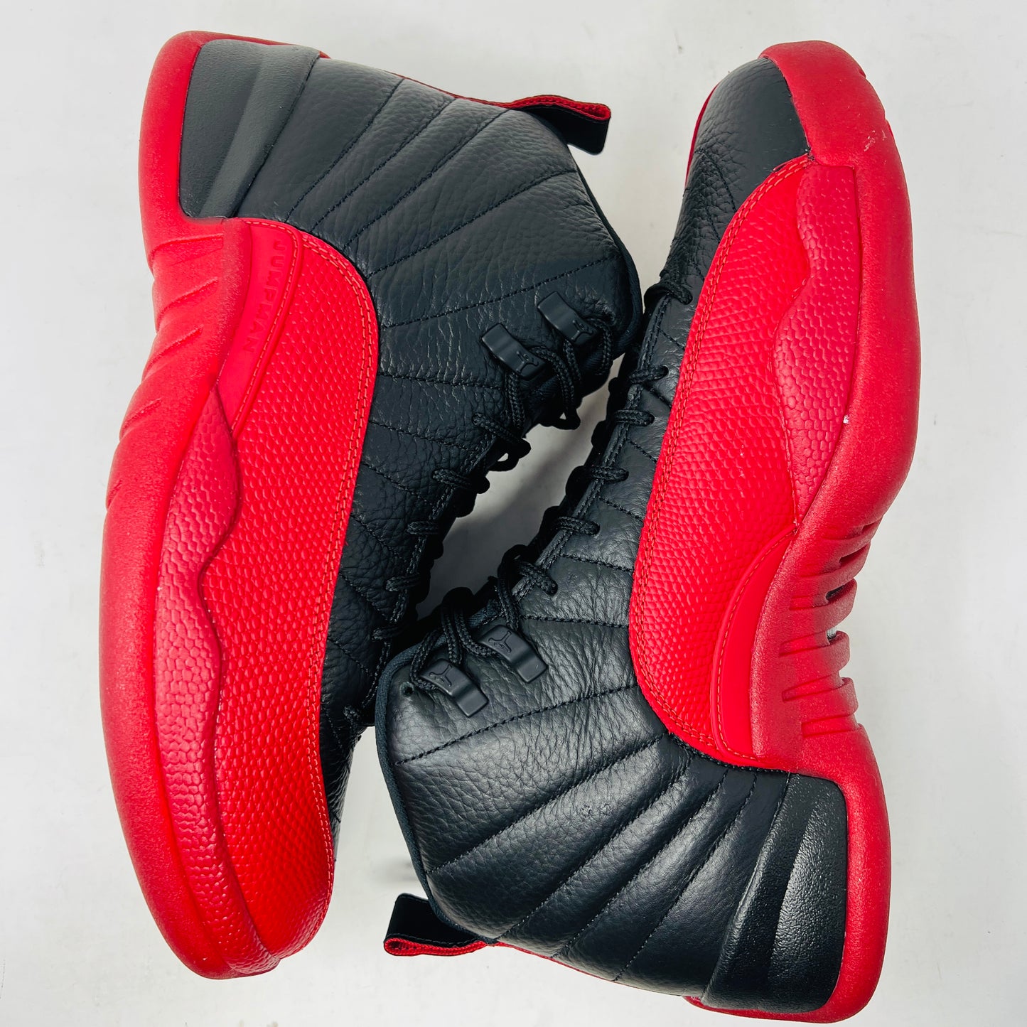 The Jordan 12 Retro Flu Game (2025) athletic shoe in black and red, brand new and pristine, sits atop an immaculate orange-and-black Nike box. It features a textured design with JUMPMAN embossed on its high-top style, perfectly enhancing this stylish accessory from the Jordan brand.