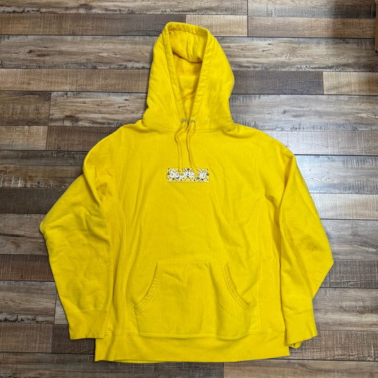 The Supreme Bandana Box Logo Hooded Sweatshirt Yellow, preowned and featuring a front pocket with a small multicolored chest graphic, is displayed laid flat on a rustic wooden floor.