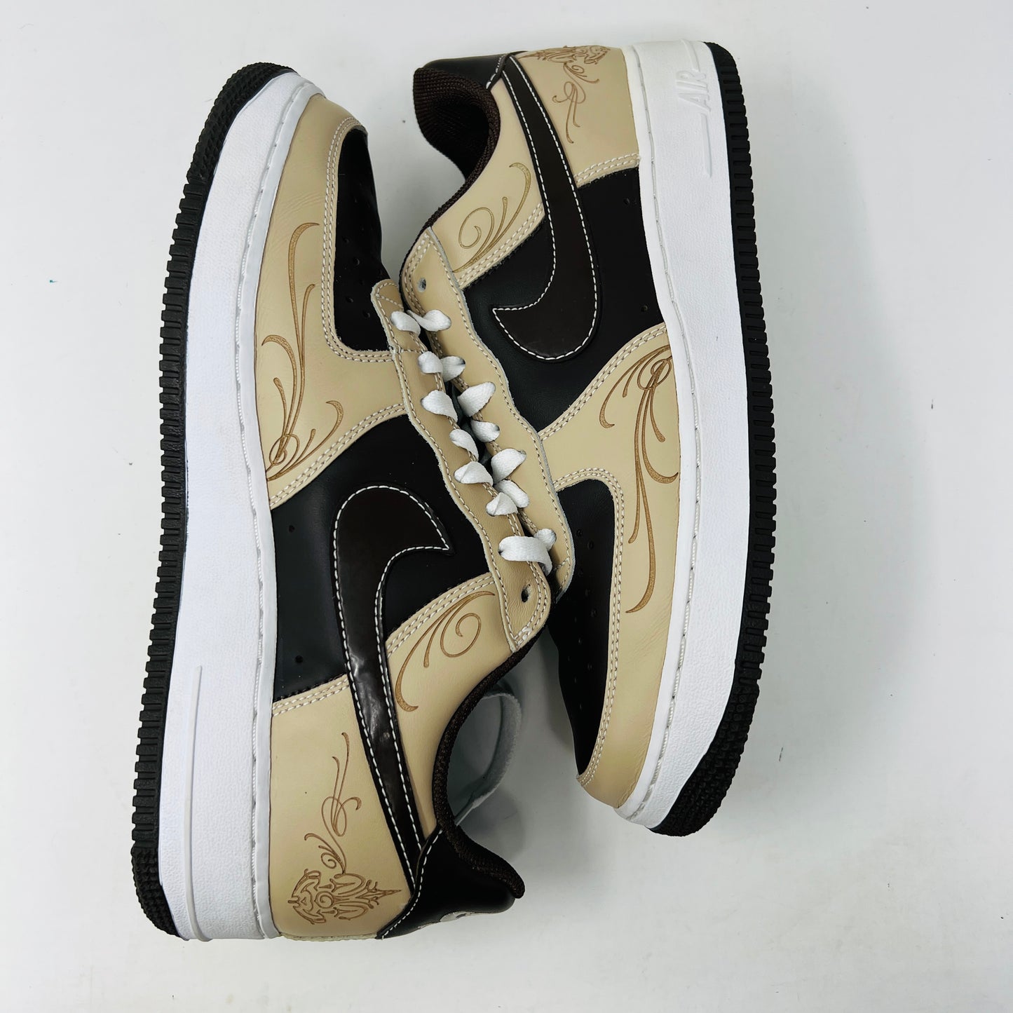 The Nike Air Force 1 Low Brown Pride sneakers feature beige and dark brown leather panels, white laces, and a white sole. They are shown from a top-down perspective in super clean condition on an immaculate white background.