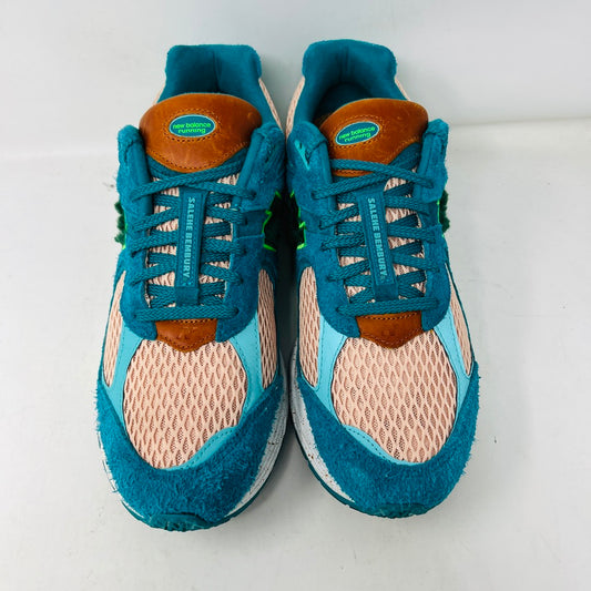 A pair of New Balance 2002R by Salehe Bembury, in colorful teal, pink, and green accents, sits atop a shoebox. Two extra lace sets—pink and green—are in front. The shoebox label details the size and style; the box is in good condition with a clean design.