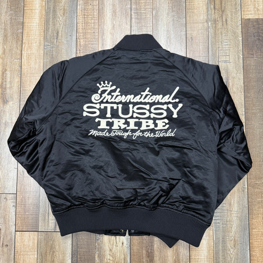 The Stussy Souvenir Jacket FW24 by Holy Ground Sneaker Shop is a black satin bomber with ribbed cuffs, collar, and hem. It features a snap button front and Stussy embroidered in white on the left chest. The jacket is laid flat on a wooden floor.