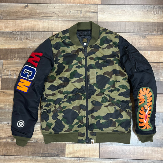 The preowned BAPE Shark MA-1 Jacket by Bape in green and brown camo features a black sleeve with the embroidered WGM letters and a cartoon tiger design, all displayed on a wooden floor.