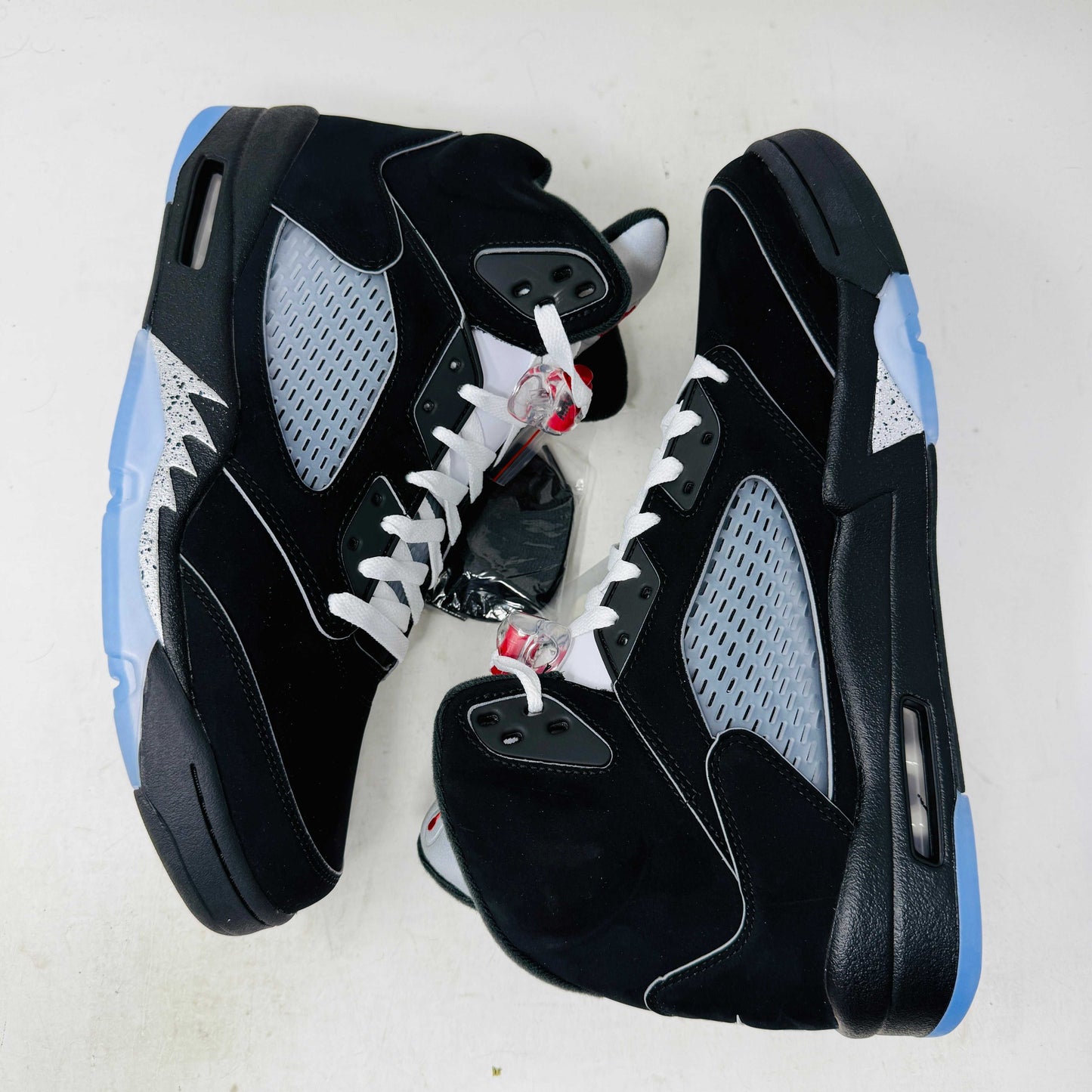 The Jordan 5 Retro OG Black Metallic Reimagined sneaker, by Jordan, comes in black with gray and silver accents, styled on a matching shoebox featuring the red 2025 Nike Air logo. This authentic shoe has a transparent sole and a signature Nike Air tag attached.