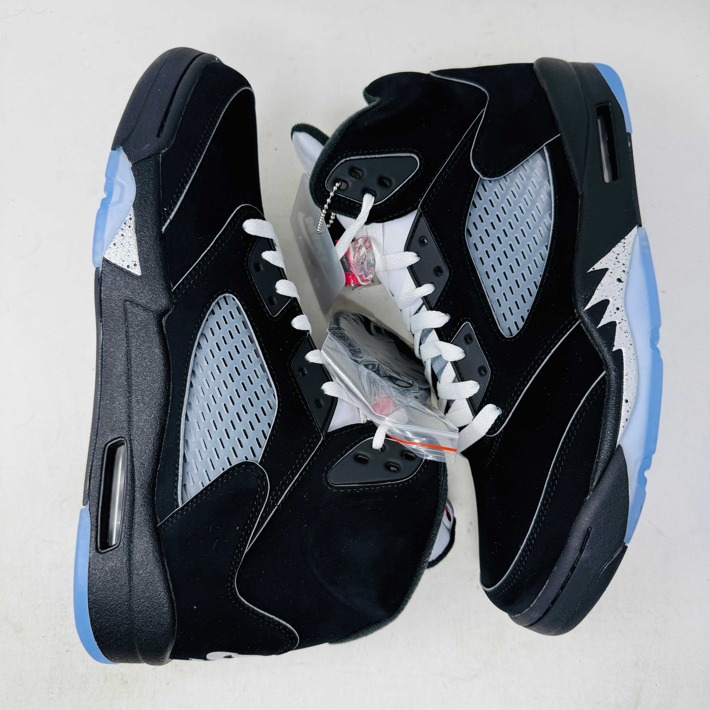 The Jordan 5 Retro OG Black Metallic Reimagined sneaker, by Jordan, comes in black with gray and silver accents, styled on a matching shoebox featuring the red 2025 Nike Air logo. This authentic shoe has a transparent sole and a signature Nike Air tag attached.