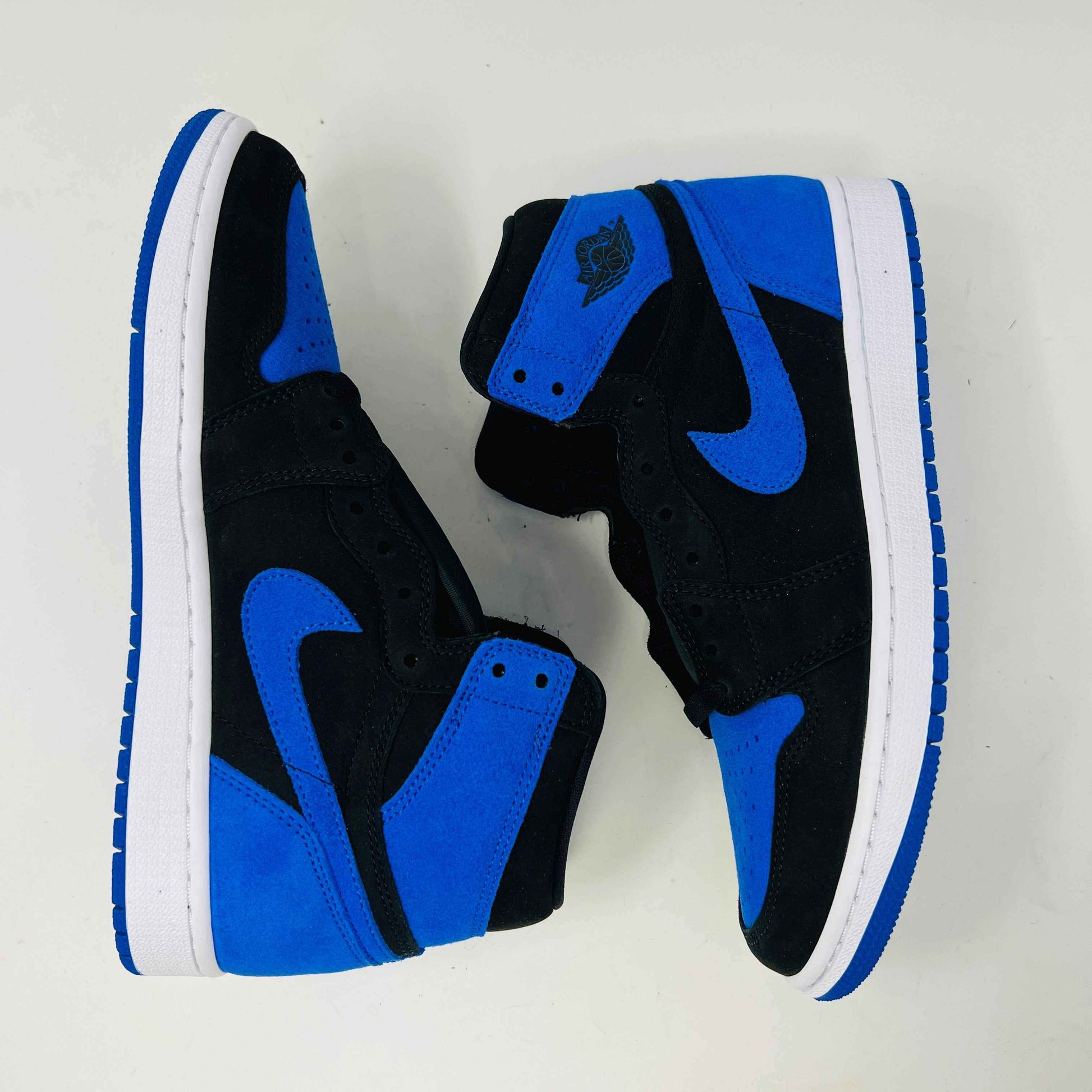 A pair of new Jordan 1 Retro High OG Royal Reimagined sneakers by Jordan rests on a matching box. These genuine shoes have blue accents on the swoosh, heel, and toe cap with a white midsole. The box also features a blue Nike logo.