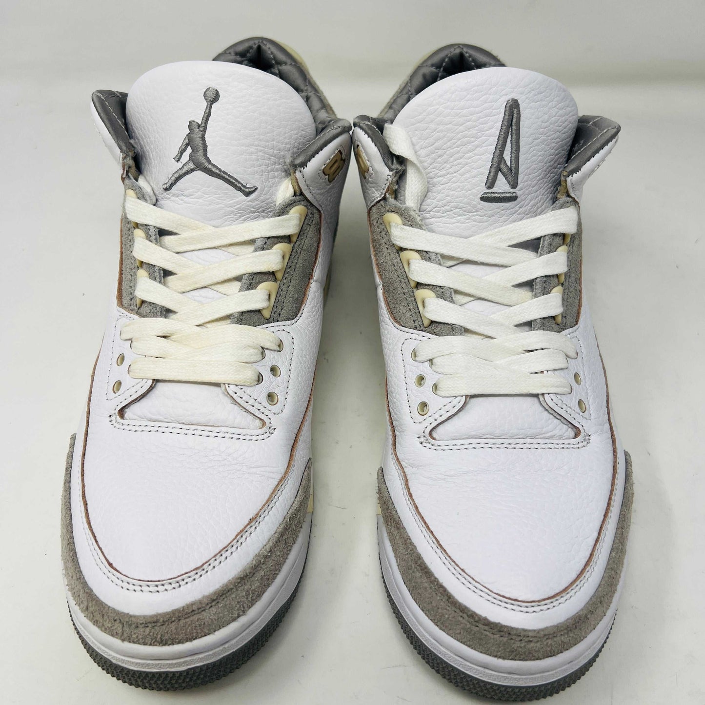 The Holy Ground Jordan 3 Retro SP A Ma Maniére (Womens) sneakers in white and gray, priced at $450, are displayed atop their box. They feature textured leather, suede accents, and include a pack of cream-colored extra laces for customization.