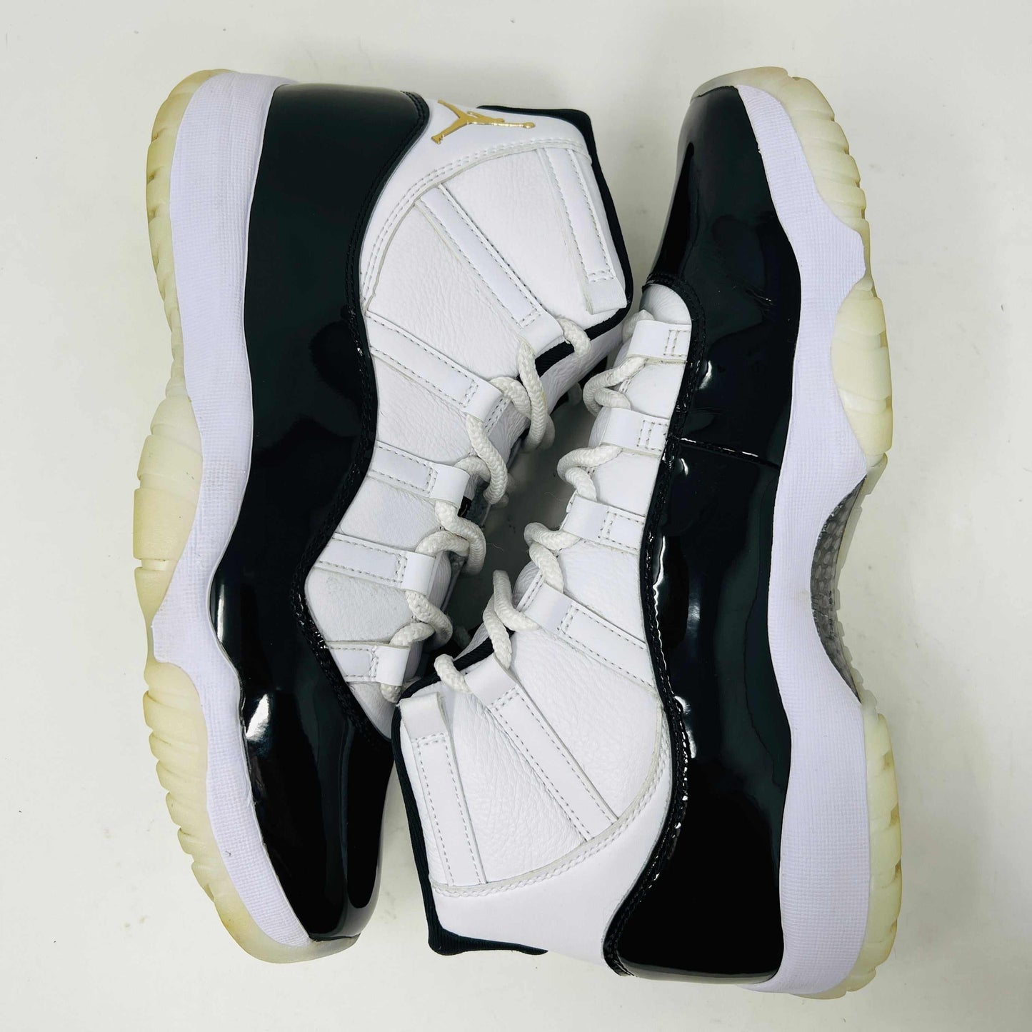 The Jordan 11 Retro DMP Gratitude (2023) sneaker, featuring white mesh, black patent leather, a translucent sole, and a gold Jumpman logo on the heel, rests elegantly on its box labeled with size and model details.