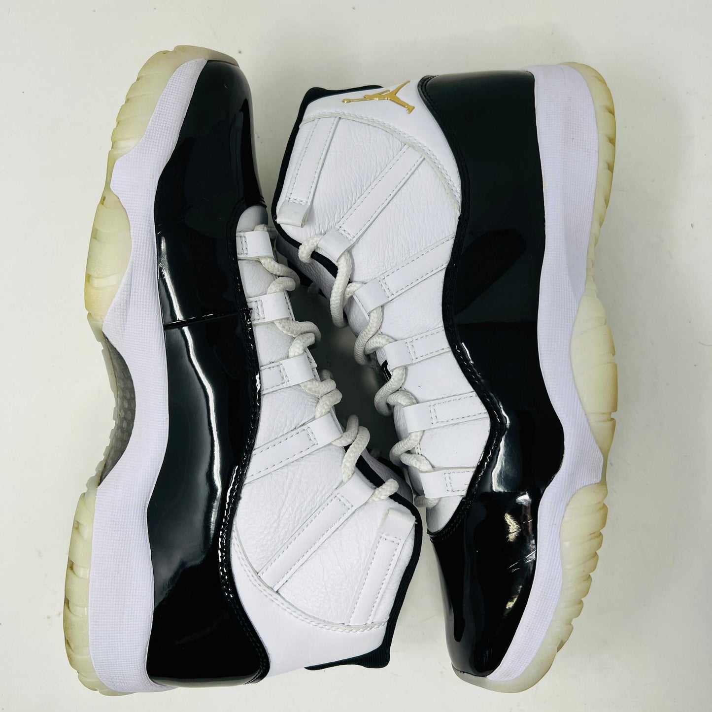 The Jordan 11 Retro DMP Gratitude (2023) sneaker, featuring white mesh, black patent leather, a translucent sole, and a gold Jumpman logo on the heel, rests elegantly on its box labeled with size and model details.