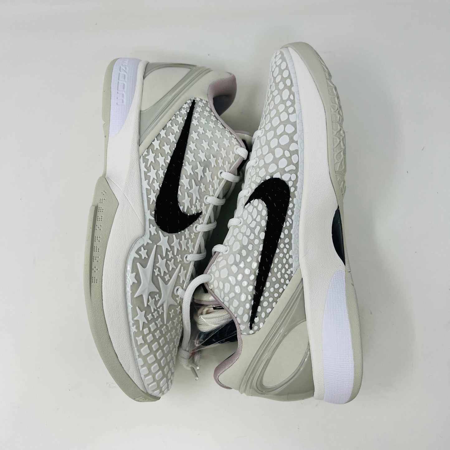 The Nike Kobe 6 Protro Sail All-Star shoes, featuring white textured patterns and black swoosh details with star designs, are elegantly displayed on a sleek black shoebox.