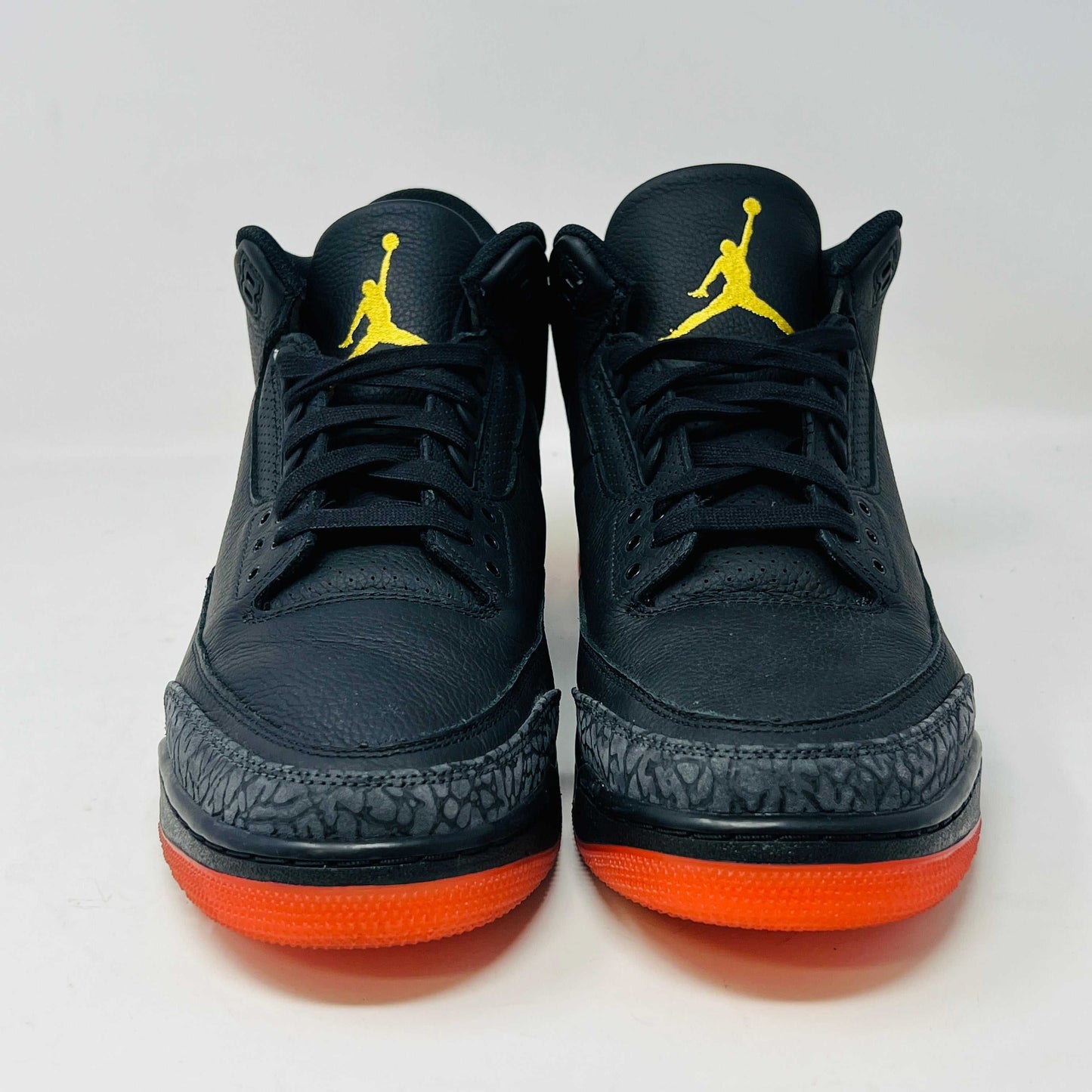 The Jordan 3 Retro J Balvin Rio sneaker in black features clean uppers and textured details with a vibrant gradient heel. It comes atop a box with a black and orange sunset design, accompanied by bright orange laces, ensuring authenticity.
