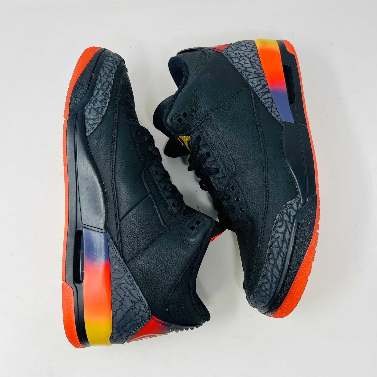 The Jordan 3 Retro J Balvin Rio sneaker in black features clean uppers and textured details with a vibrant gradient heel. It comes atop a box with a black and orange sunset design, accompanied by bright orange laces, ensuring authenticity.
