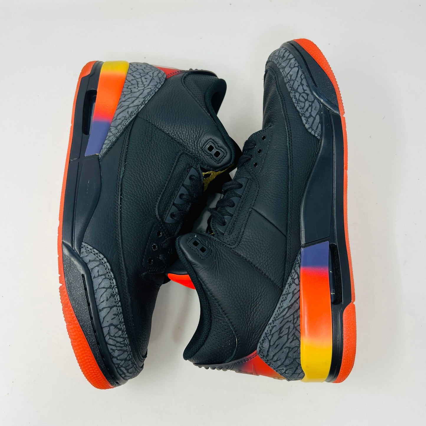The Jordan 3 Retro J Balvin Rio sneaker in black features clean uppers and textured details with a vibrant gradient heel. It comes atop a box with a black and orange sunset design, accompanied by bright orange laces, ensuring authenticity.