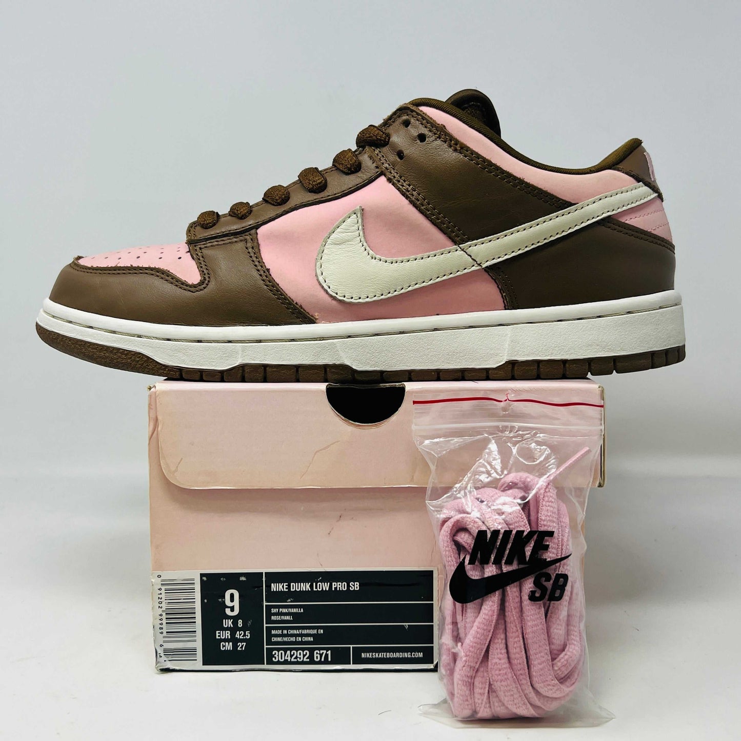 A 2005 Nike SB Dunk Low Stussy Cherry in brown and pink rests on its box, featuring a cream swoosh. The authentic shoe includes extra pink laces in a clear NIKE SB bag. The box label shows size 9.
