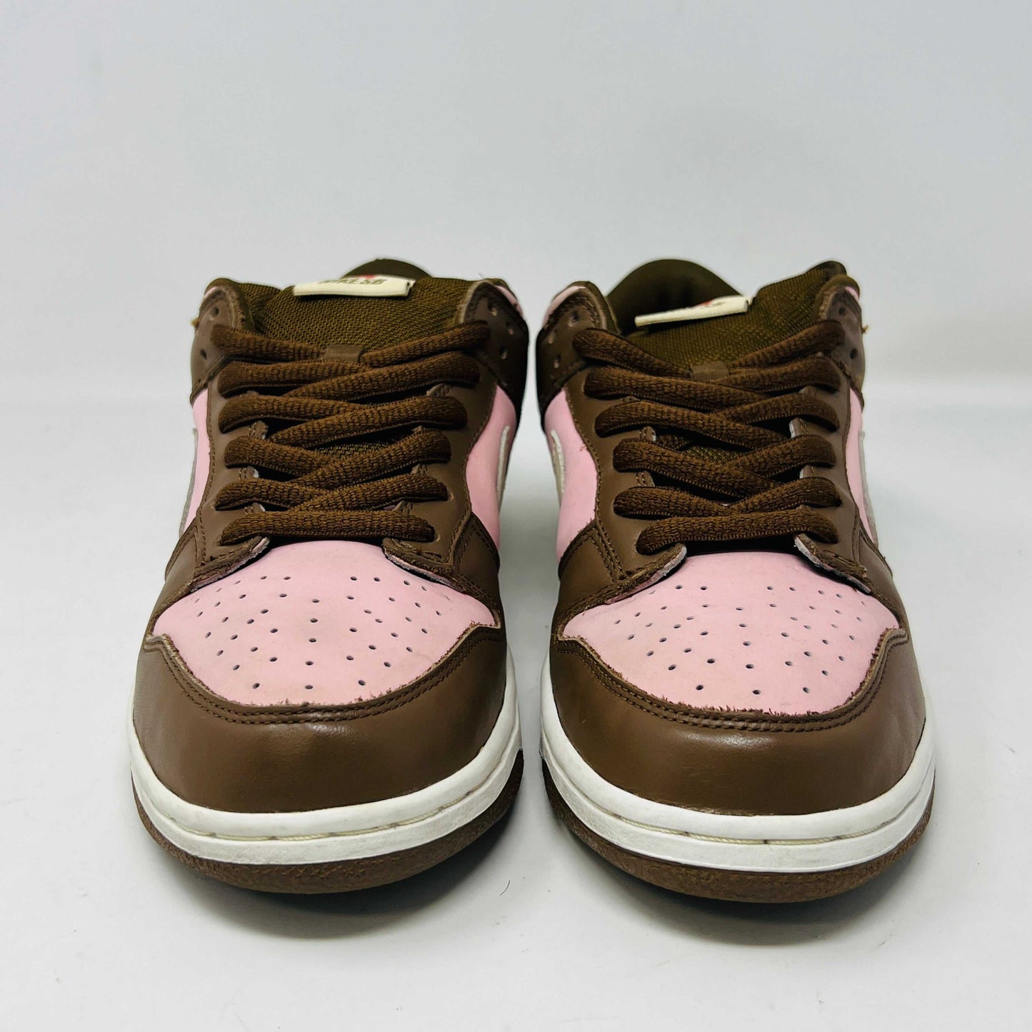 A 2005 Nike SB Dunk Low Stussy Cherry in brown and pink rests on its box, featuring a cream swoosh. The authentic shoe includes extra pink laces in a clear NIKE SB bag. The box label shows size 9.