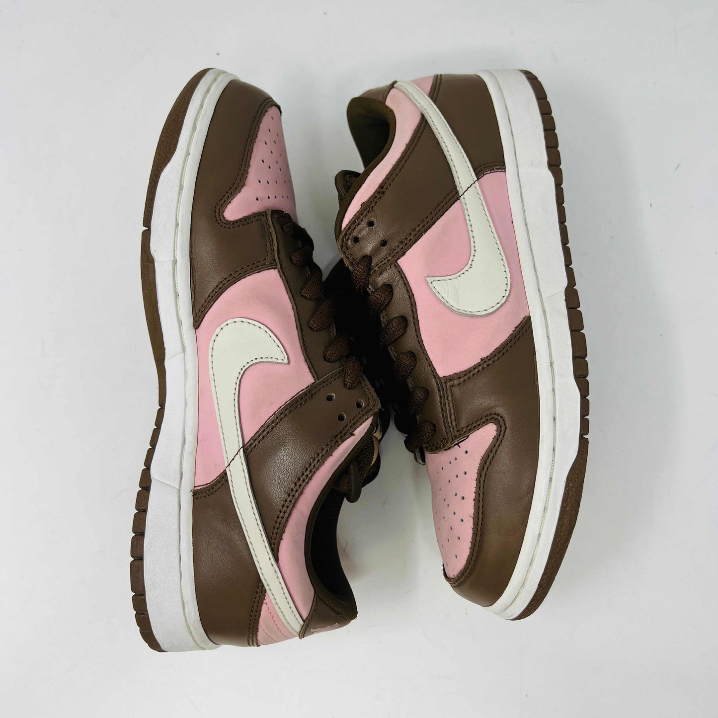A 2005 Nike SB Dunk Low Stussy Cherry in brown and pink rests on its box, featuring a cream swoosh. The authentic shoe includes extra pink laces in a clear NIKE SB bag. The box label shows size 9.