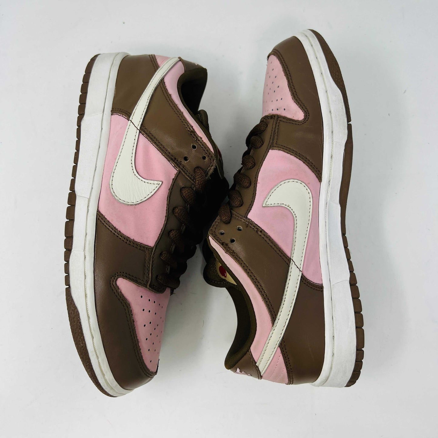 A 2005 Nike SB Dunk Low Stussy Cherry in brown and pink rests on its box, featuring a cream swoosh. The authentic shoe includes extra pink laces in a clear NIKE SB bag. The box label shows size 9.