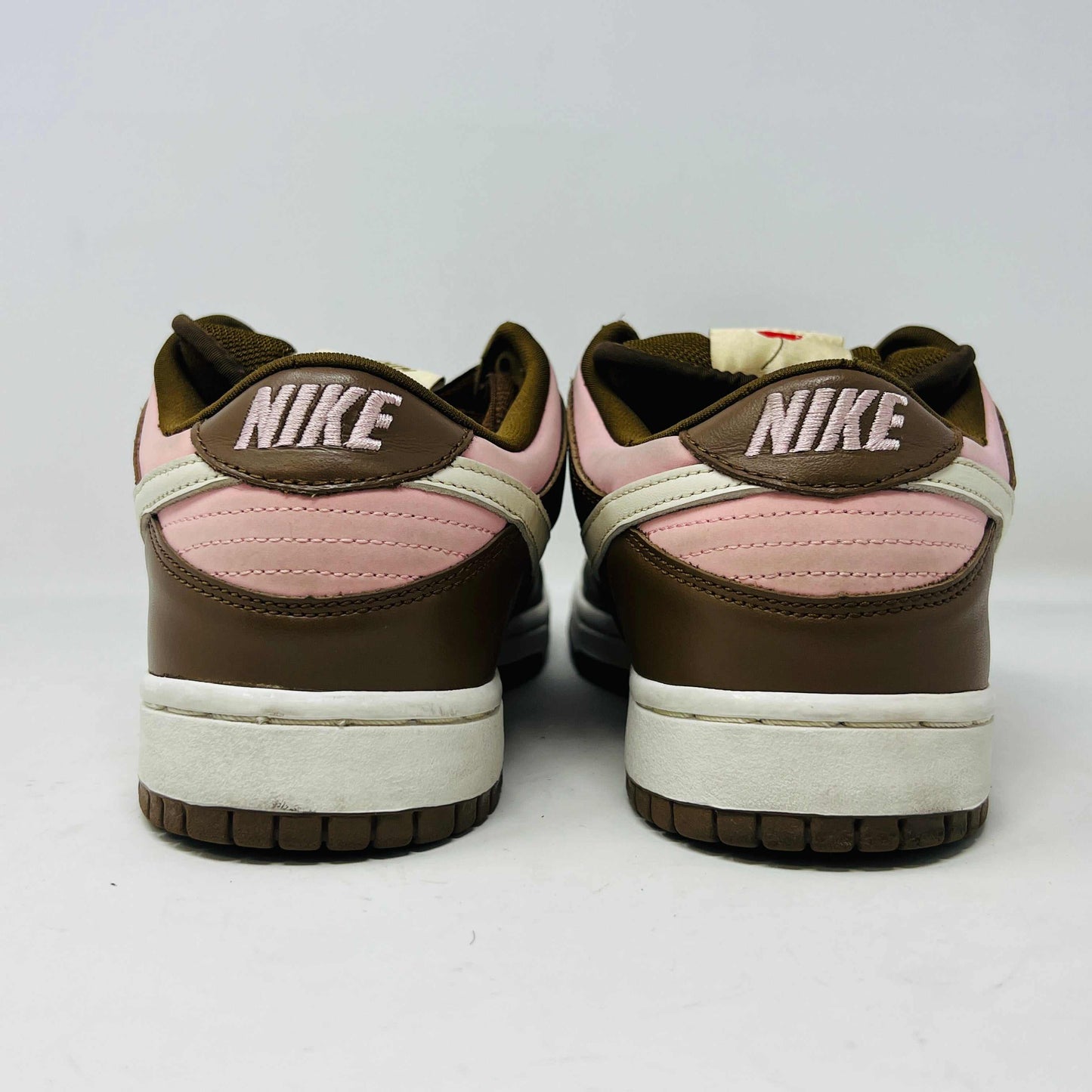 A 2005 Nike SB Dunk Low Stussy Cherry in brown and pink rests on its box, featuring a cream swoosh. The authentic shoe includes extra pink laces in a clear NIKE SB bag. The box label shows size 9.
