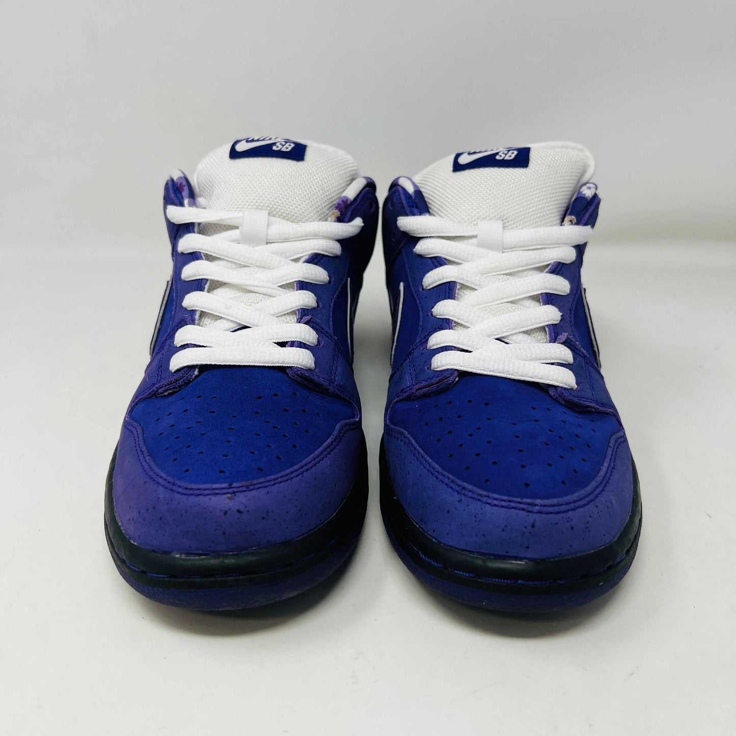 The Nike SB Dunk Low Concepts Purple Lobster sneaker, featuring a clean upper with a white swoosh, rests on a blue and white Nike SB shoebox. An extra bag with pink laces adds a vibrant touch in front of the box.