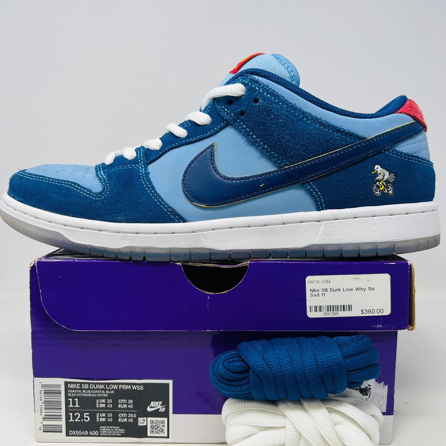 A Nike SB Dunk Low Pro Why So Sad? shoe, with blue uppers and white sole, rests on a purple box. It features an embroidered graphic on the heel, a dark blue swoosh, and extra laces in front. Price tags from 2022 are visible beside it.