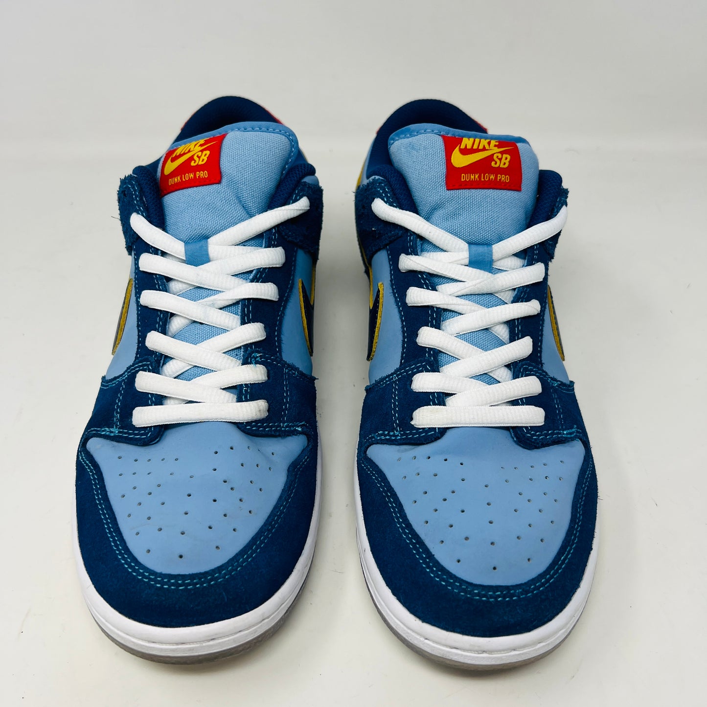 A Nike SB Dunk Low Pro Why So Sad? shoe, with blue uppers and white sole, rests on a purple box. It features an embroidered graphic on the heel, a dark blue swoosh, and extra laces in front. Price tags from 2022 are visible beside it.