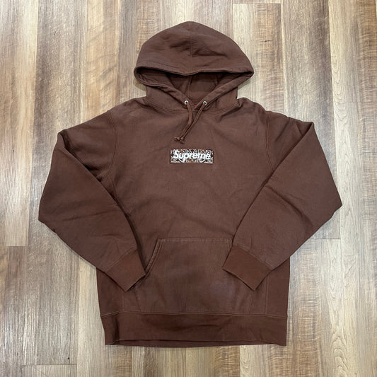 The Supreme Bandana Box Logo Hooded Sweatshirt in Dark Brown is preowned, featuring a kangaroo pocket, drawstrings, and a chest logo patch. Displayed on an earthy-toned wooden floor, it exudes timeless appeal.