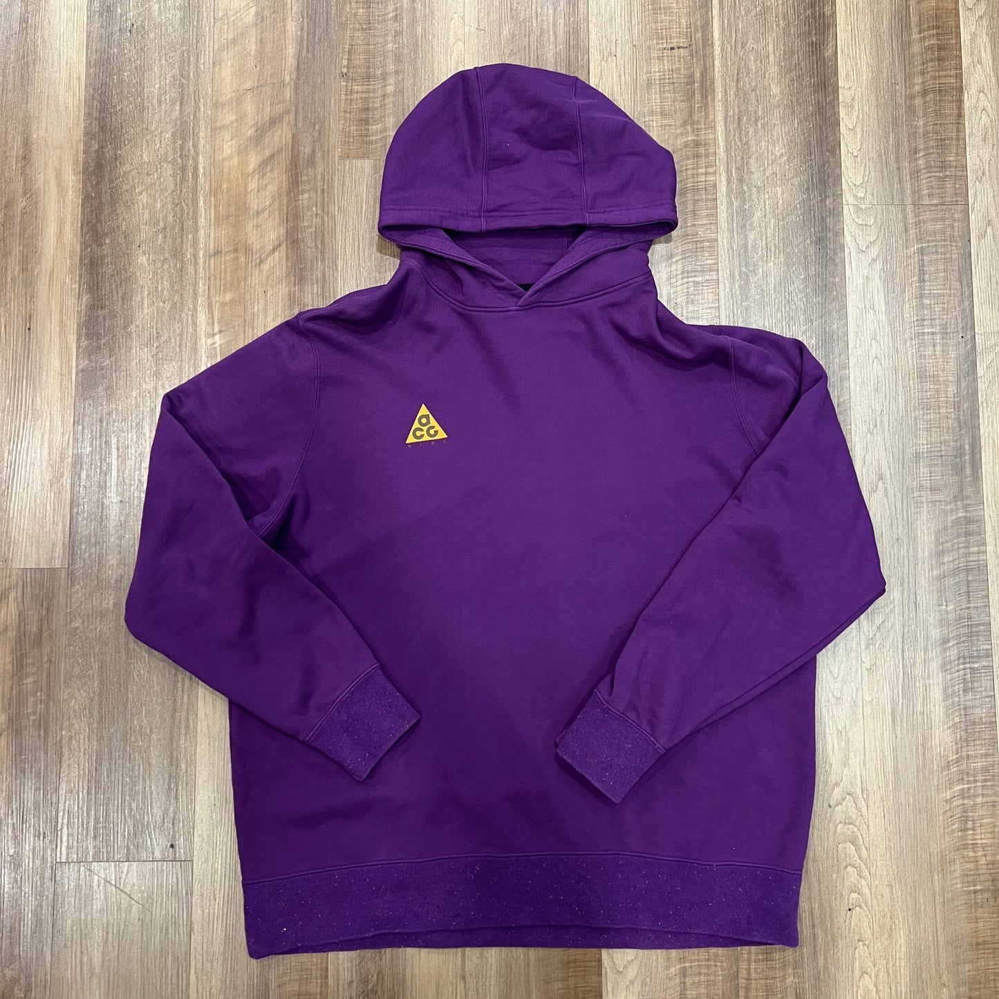 A preowned Nike ACG Hoodie Purple is placed on a wooden floor. It features long sleeves, a front pocket, and displays a small yellow ACG triangle logo on the chest.